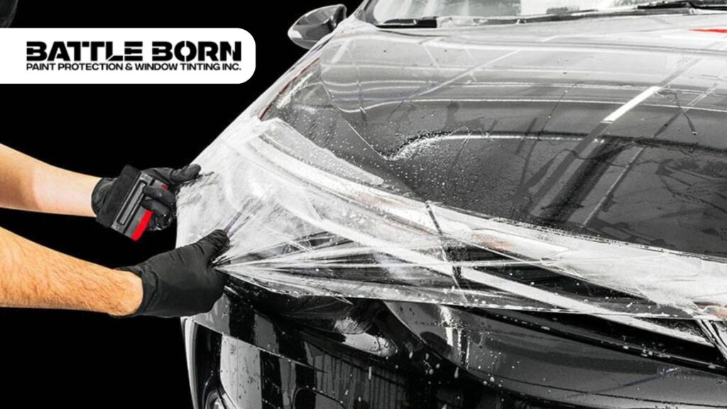 A technician wearing black gloves carefully applies a transparent paint protection film (PPF) to the front of a black car, using a squeegee to smooth out the surface. The Battle Born Paint Protection & Window Tinting Inc. logo is visible in the top left corner