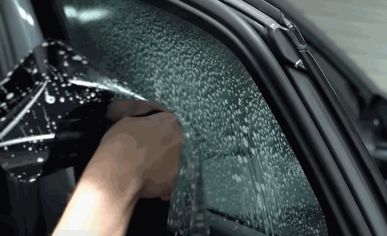 A hand applies window tint film to a wet car window, smoothing it over the glass.