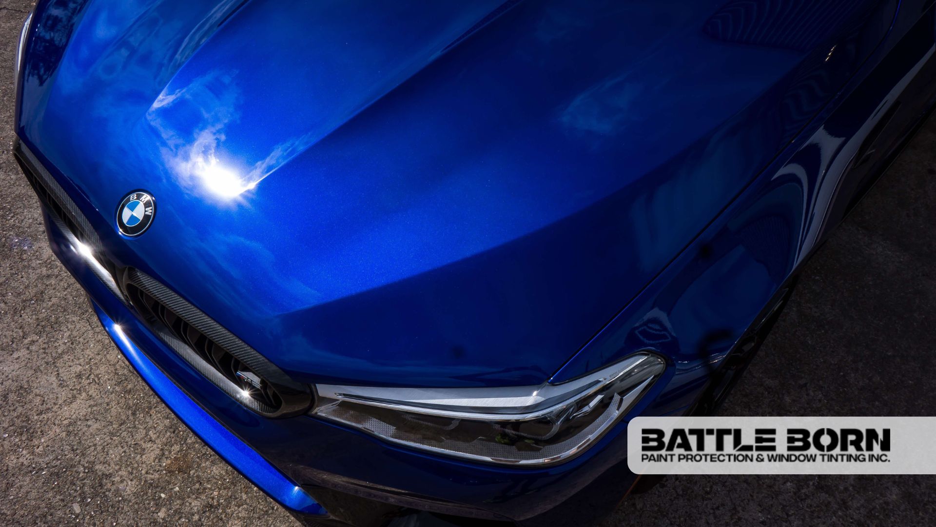 Close-up of a glossy blue BMW hood reflecting the sky, highlighting a sleek headlight and the BMW emblem