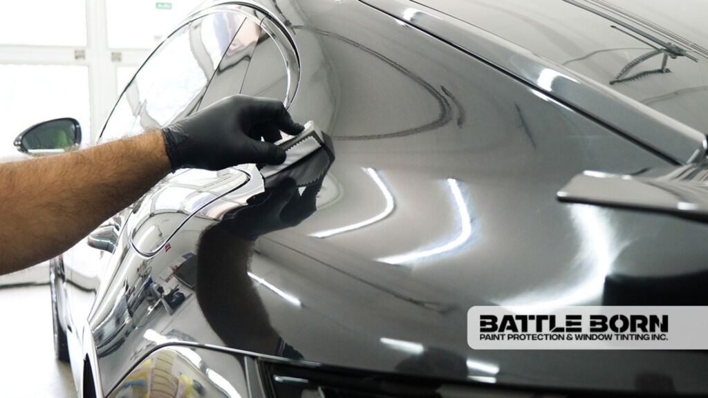 A detailer applies ceramic coating to the rear panel of a black car using an applicator pad, with reflections of the garage visible in the glossy finish.