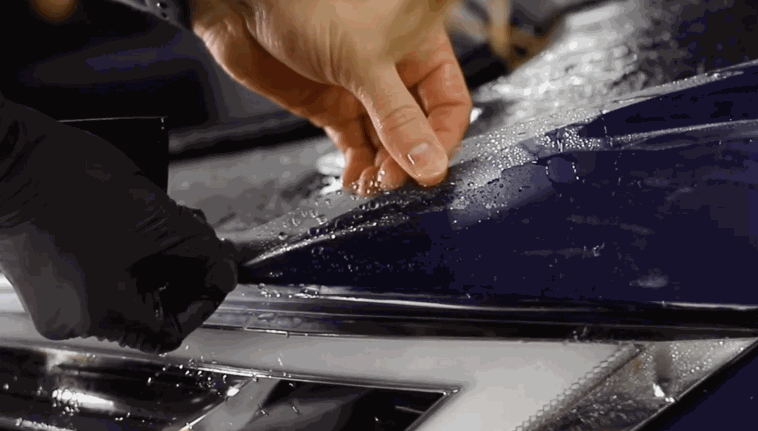 A person aligns and presses paint protection film onto a wet blue vehicle surface, securing it along the headlight edge.