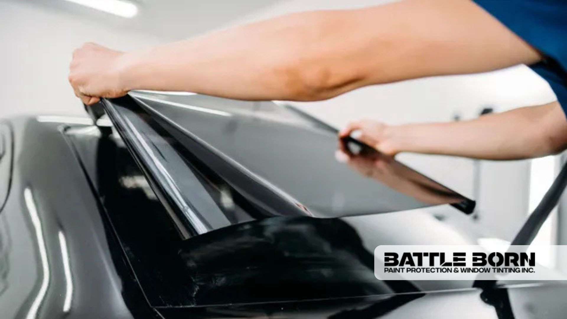 Close-up of a technician applying dark window tint film to the rear windshield of a black vehicle. The image includes the Battle Born Paint Protection & Window Tinting Inc. logo.