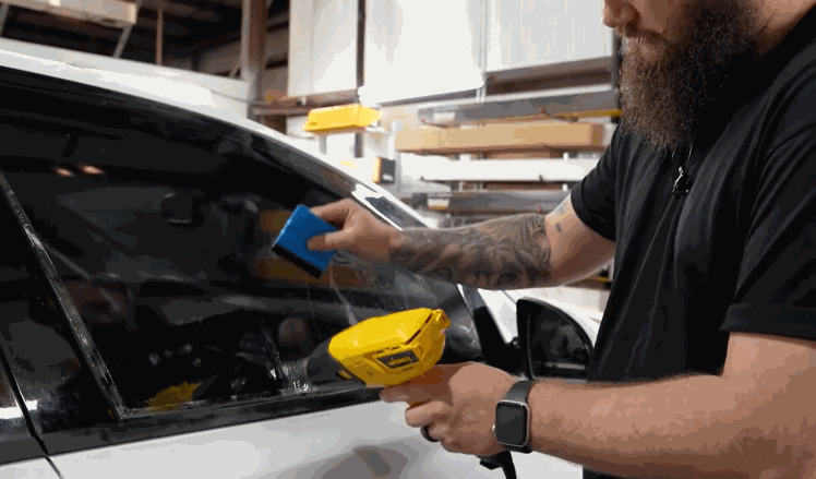 A bearded technician uses a heat gun and squeegee tool to shape and seal window tint film onto a car window.