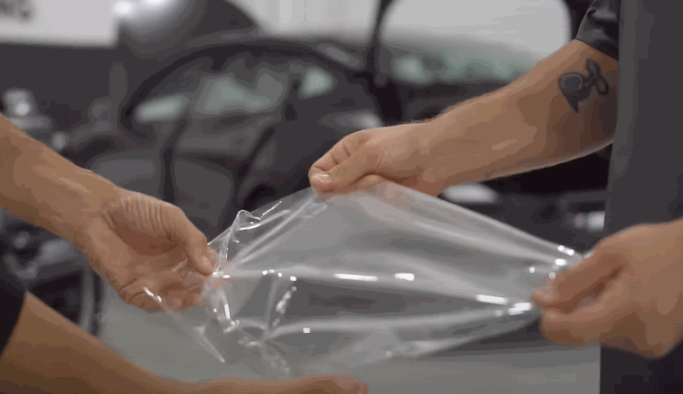 Two people stretch a sheet of clear protective film before applying it to a vehicle surface.