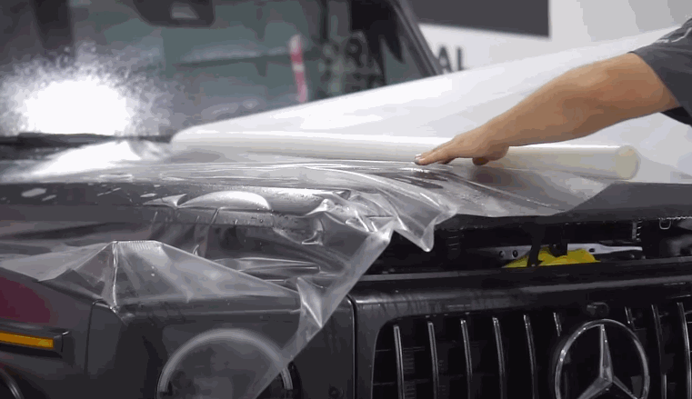A technician smoothly applies a large clear paint protection film to the hood of a black Mercedes-Benz SUV.