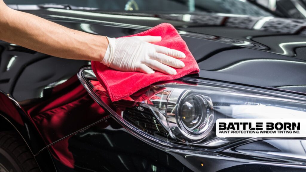 A person wearing white gloves buffs a car headlight and hood with a red microfiber cloth to apply or remove wax.