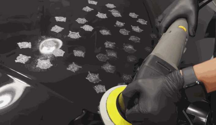 White dabs of polish or wax are applied evenly across a black car hood before being buffed in with a polishing machine.