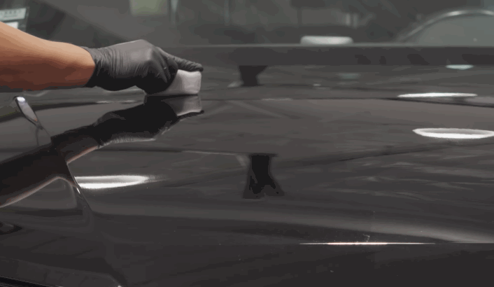 A gloved hand applies ceramic coating to a black car hood using a soft applicator pad, creating a sleek, protective finish.