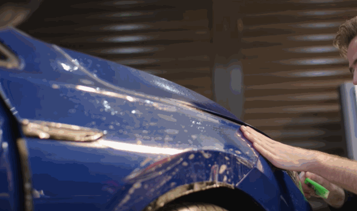Technician pressing a blue car fender with a squeegee to lay down paint protection film.