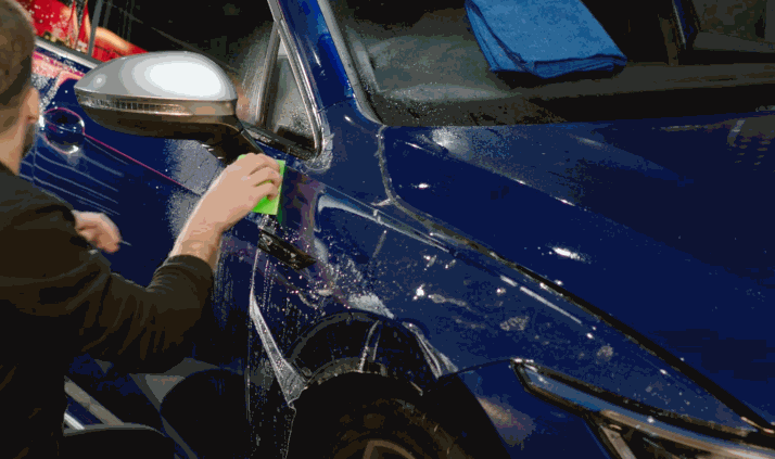 Installer using a green squeegee to smooth paint protection film onto the front side panel of a blue car.