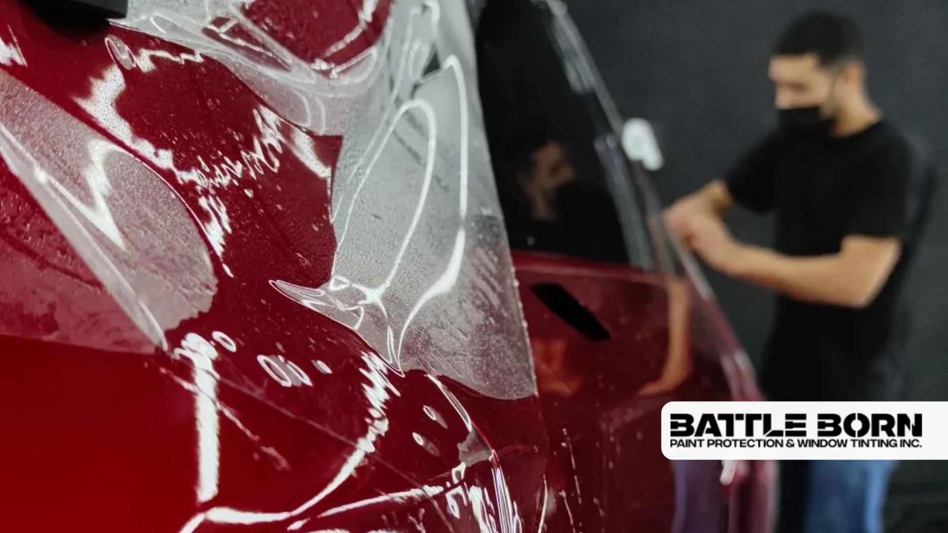 Close-up of wet paint protection film being applied to a red car, with an installer working in the background