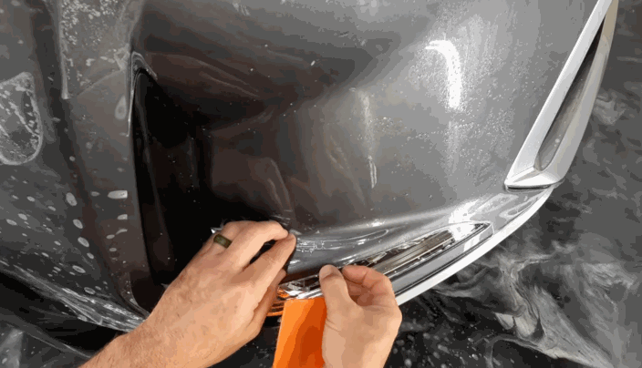 Hands using an orange tool to press paint protection film around the curve of a car’s bumper, ensuring it conforms smoothly to the surface.
