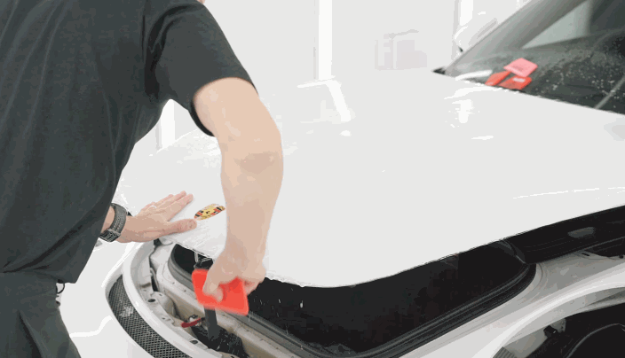 A technician installs paint protection film onto the hood of a white Porsche, using a red squeegee to remove excess moisture and air.
