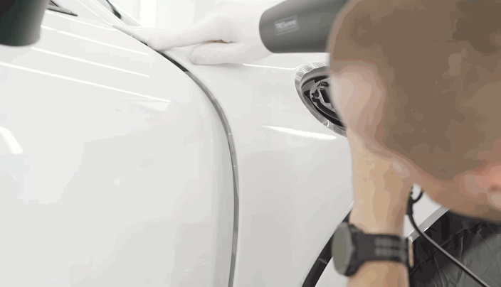 A detailer uses a heat gun to stretch and adhere paint protection film onto a car’s side panel, working near the edge of the gas cap.