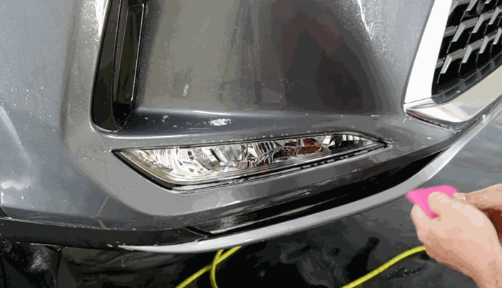 Close-up of a person applying paint protection film to a wet car bumper using a pink squeegee, carefully smoothing out air bubbles around the fog light.