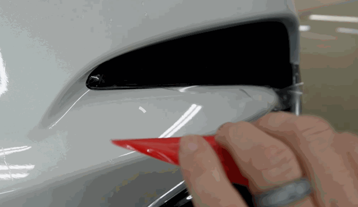 A close-up of a hand using a red squeegee to smooth paint protection film onto a white car’s bumper.