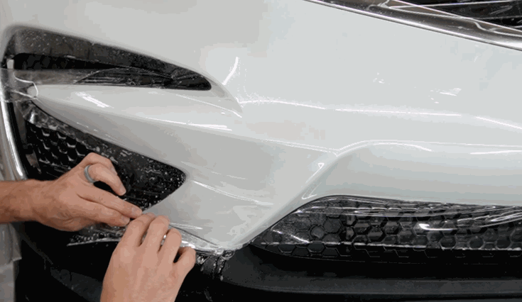 A technician presses and smooths transparent paint protection film onto the front bumper and grille of a white car.