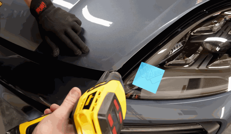 A technician uses a heat gun to apply paint protection film to the edge of a car’s hood while another person steadies the panel with gloved hands.