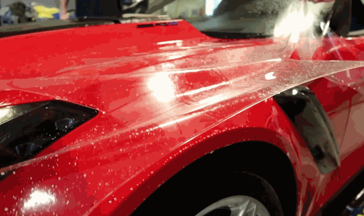 Clear paint protection film being applied smoothly over the hood of a red sports car, highlighting water beading during installation.