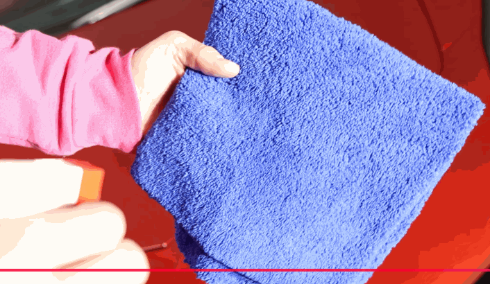 Person in a pink sleeve holding a bright blue microfiber towel over a red car surface while preparing to wipe off ceramic coating residue.