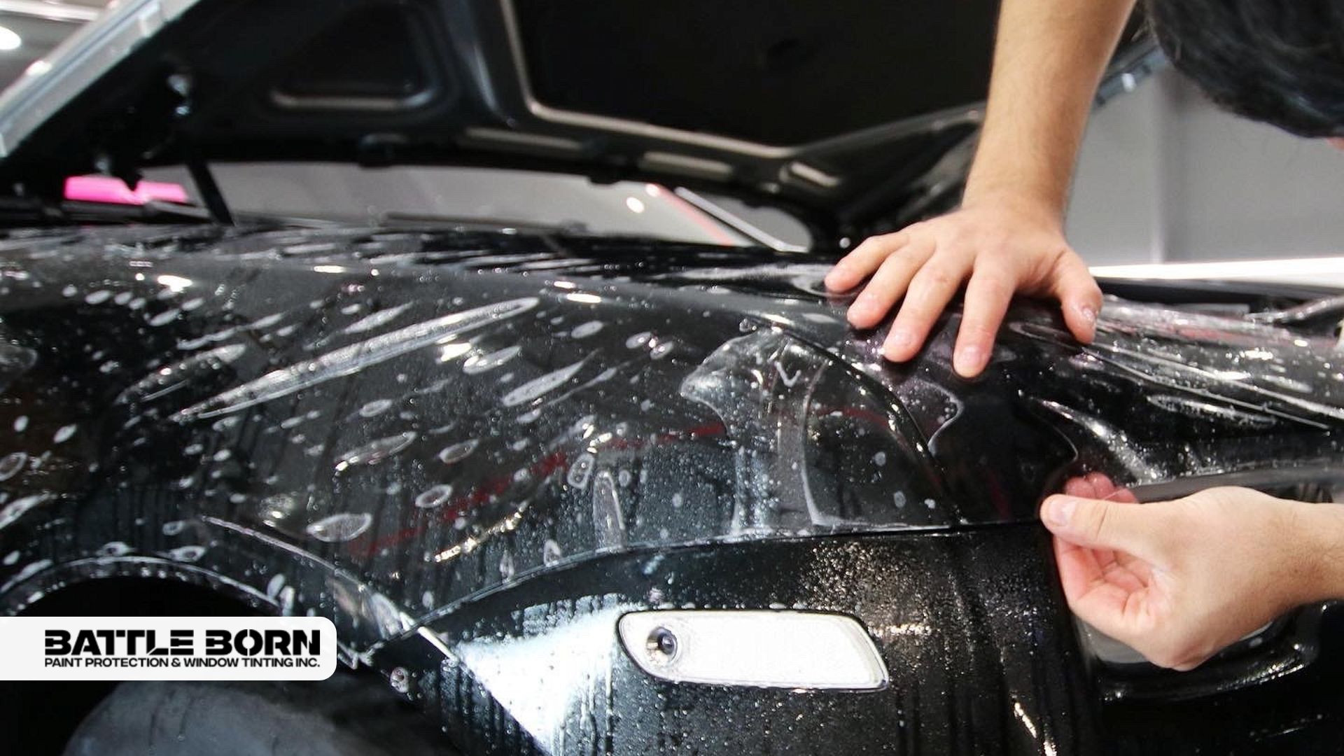Technician carefully smooths out paint protection film on a wet black vehicle hood, ensuring a bubble-free finish