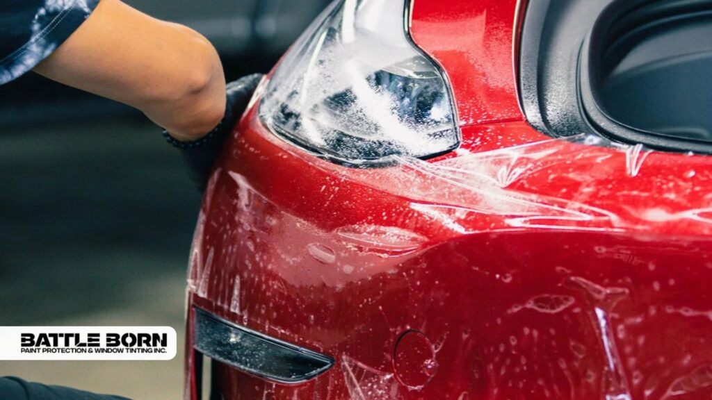 Technician working on the replacement of paint protection film around the headlight of a red car, with film partially applied and water solution visible.