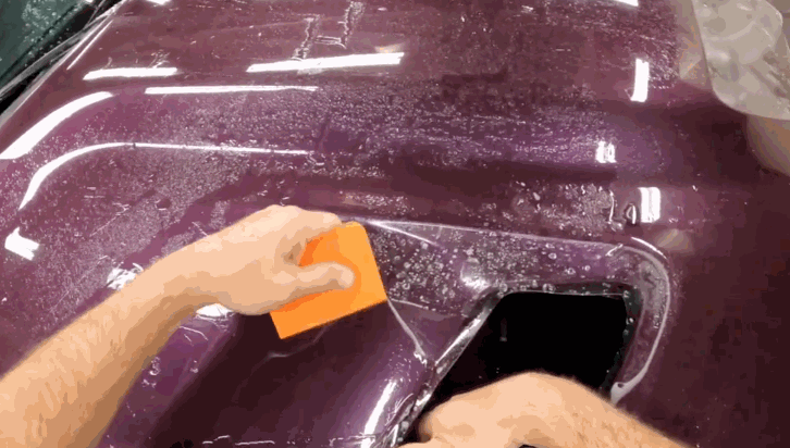 Technician presses an orange squeegee over paint protection film on a maroon bumper to remove air and water during installation. The glossy surface reflects overhead lights as the film is secured in place.
