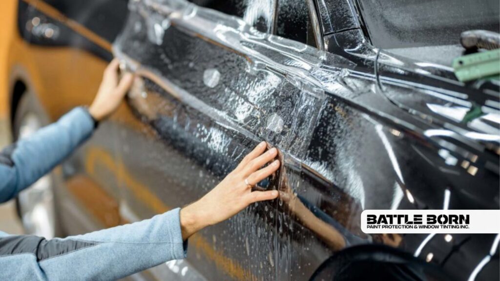 Installer aligns and smooths paint protection film across the side of a black vehicle with water solution visible beneath the surface. The film is positioned carefully along the door and trim for full coverage.