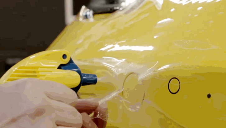 Installer uses a heat gun while stretching paint protection film over a yellow car bumper to conform it around a circular sensor. The clear film is carefully shaped to fit tight curves and edges.