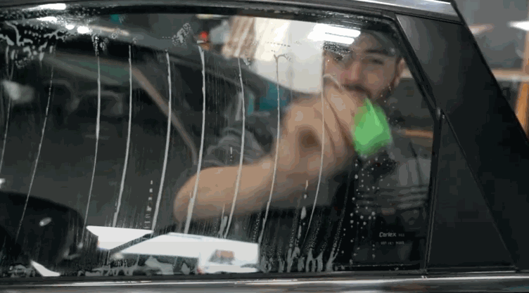 A worker wipes and smooths a car window covered in soapy solution while installing window tint from the inside of the vehicle. Vertical streaks of liquid run down the glass as the tint is positioned and secured.