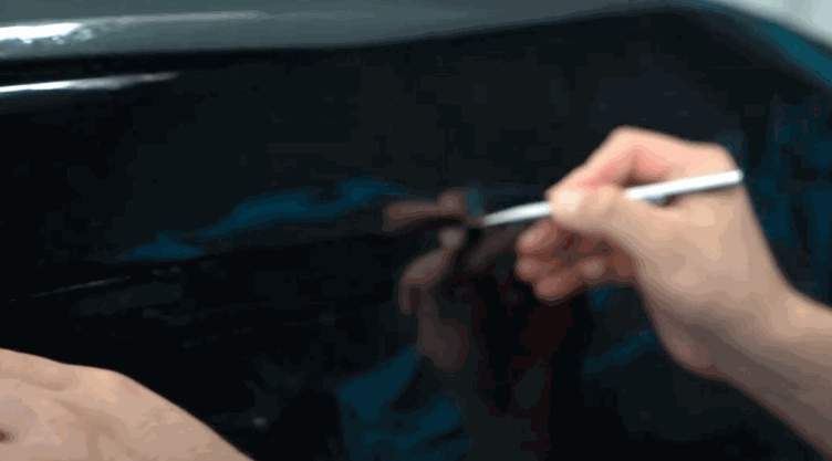 A technician carefully uses a small tool to smooth the edge of freshly applied window tint on a car window. The close up view shows the dark film being pressed flat against the glass to remove bubbles and ensure a clean finish.