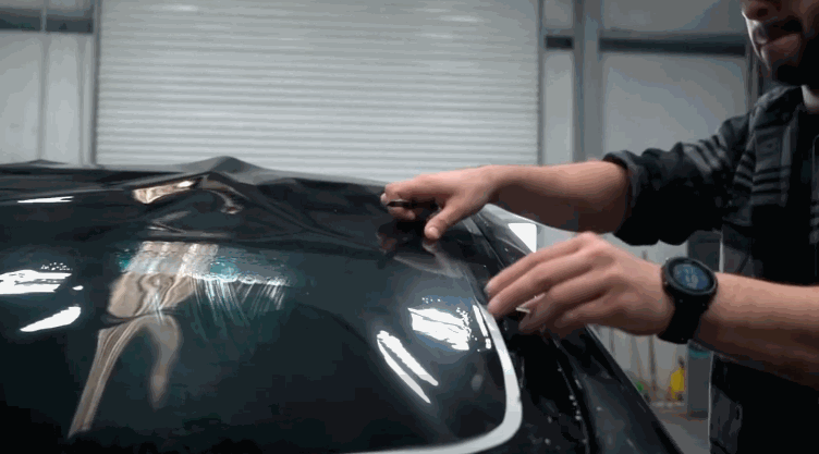 Technician stretches and smooths window tint film across a car hood surface, removing wrinkles and air pockets. The reflective surface shows light patterns as the film is adjusted into place. This demonstrates the careful handling required during window tint application.