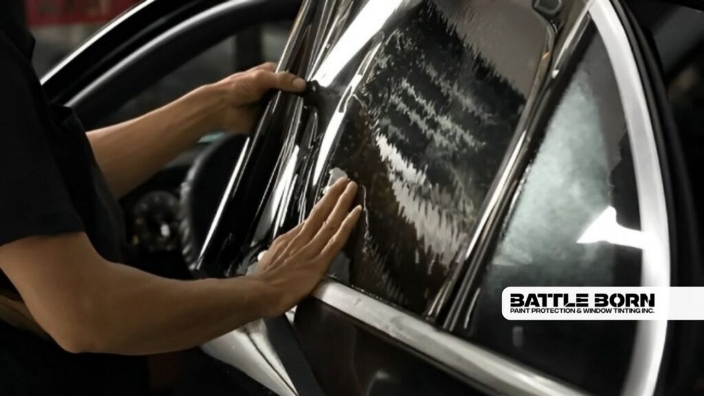 Technician applies window tint film to a car side window using both hands to smooth out bubbles and water lines. Reflections ripple across the dark film as it is carefully aligned.