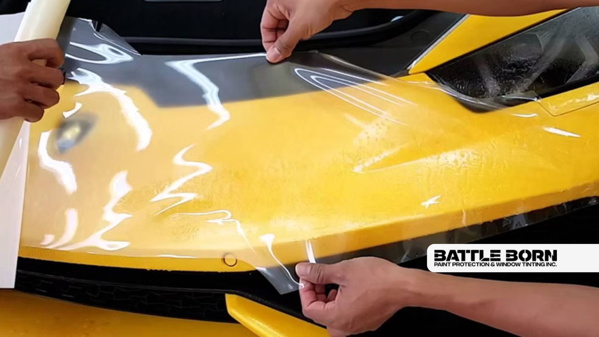 Hands carefully apply a clear paint protection film across the front of a bright yellow car hood to prevent scratches and damage. The glossy surface reflects light as the film is smoothed into place near the headlight.